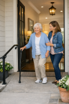 Senior woman entering her home with support from her adult daughter, using a cane and handrail on a gently sloped, no-step entryway with soft lighting and a wide, clear path.